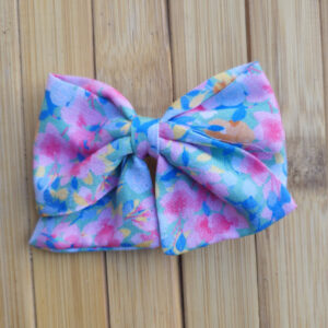 Lazo infantil de tela con estampado floral en tonos pastel azul, rosa, amarillo y verde sobre fondo de madera.