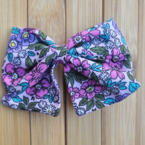 Lazo infantil estampado con flores en tonos rosa, azul y verde sobre fondo de madera.