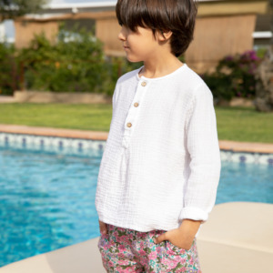 Niño con camisa blanca y pantalones cortos florales, posando junto a la piscina.