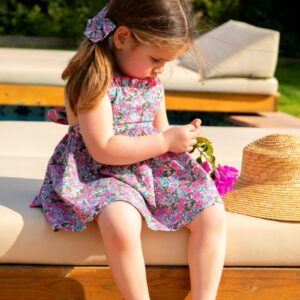 Niña pequeña sentada junto a la piscina con vestido floral de la colección Cumbia y lazo a juego, sosteniendo una flor.