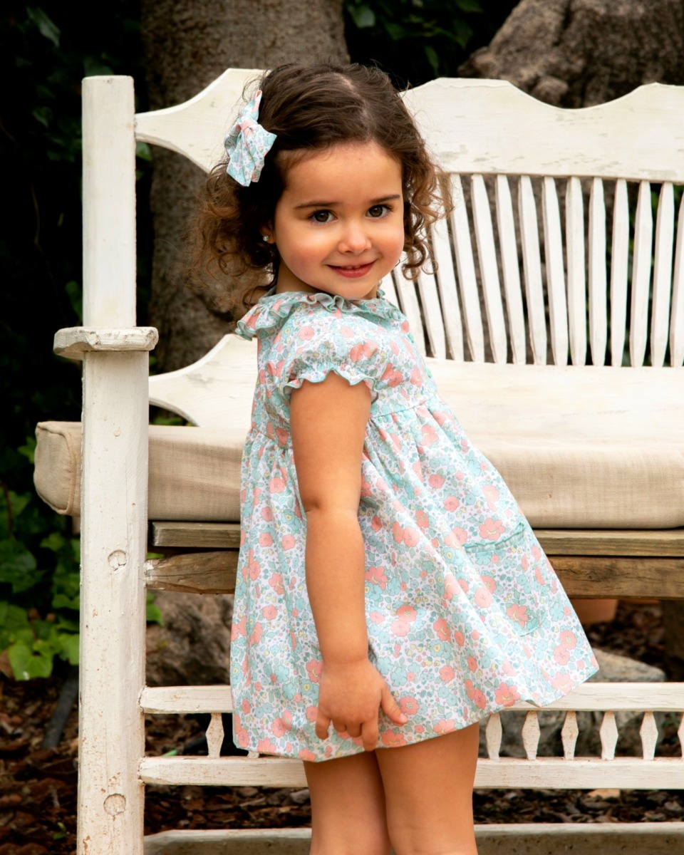 Niña pequeña con vestido floral en tonos azul y rosa y lazo a juego posa sonriente frente a un banco de madera, luciendo un conjunto de la colección Samba.