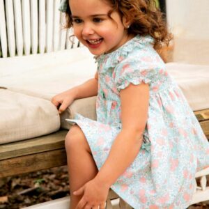 Niña sonriendo mientras lleva un vestido floral de la colección Samba, jugando y sentada en un banco de madera.