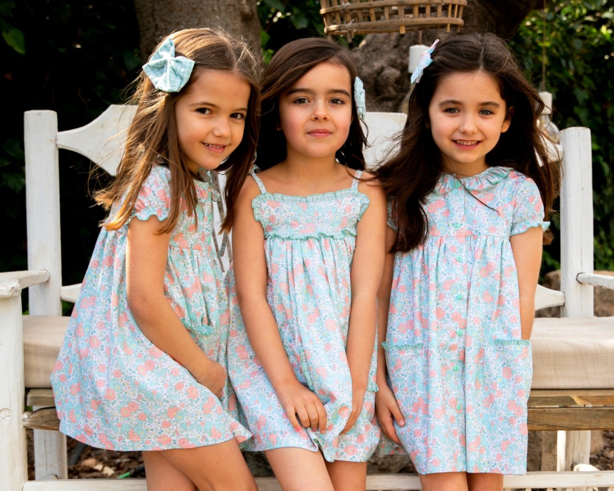 Tres niñas sonrientes usando vestidos florales de la colección Samba, posando juntas en un banco de madera.