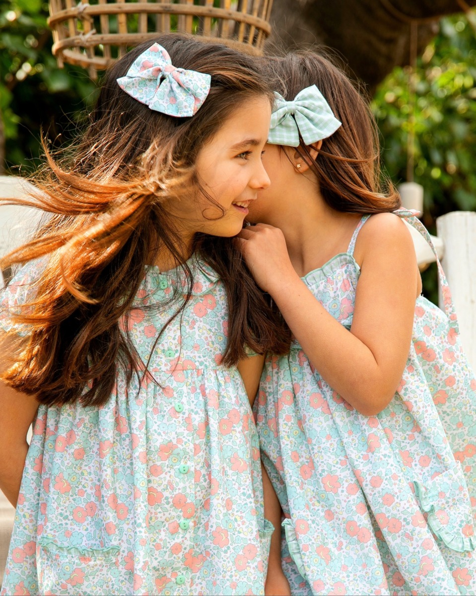 Niñas usando vestidos de flores con lazo, compartiendo un momento de complicidad, mostrando el diseño colorido y fresco de la colección.