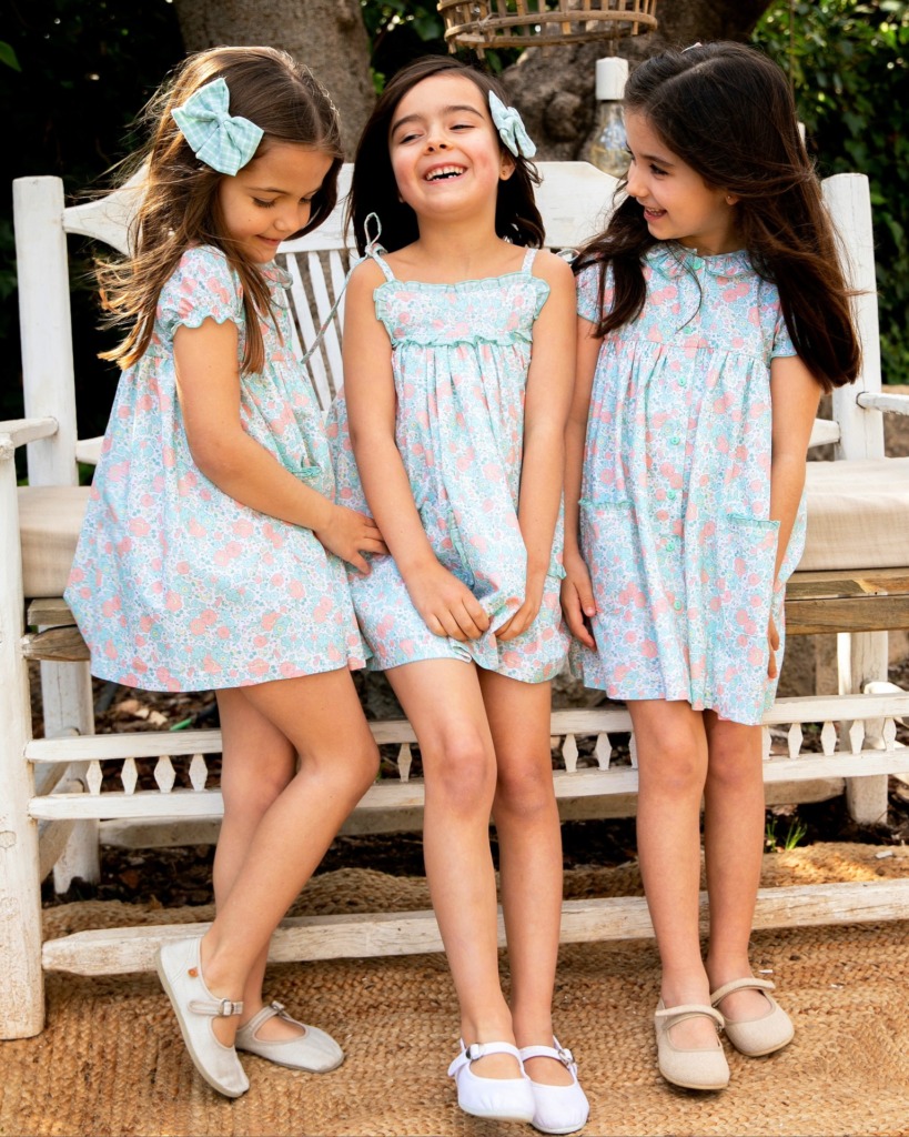 Imagen actual: Tres niñas con vestidos florales y lazos, sentadas al aire libre en primavera.