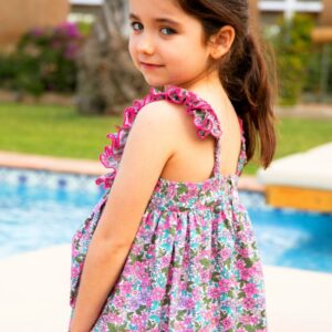 Niña con vestido floral de tirantes y volantes fucsias, de espaldas, girando la mirada junto a una piscina. Colección Cumbia.