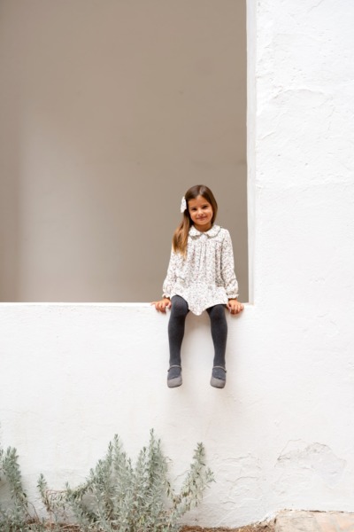 Niña sentada en un muro blanco con vestido floral y leotardos grises, moda infantil Sutier