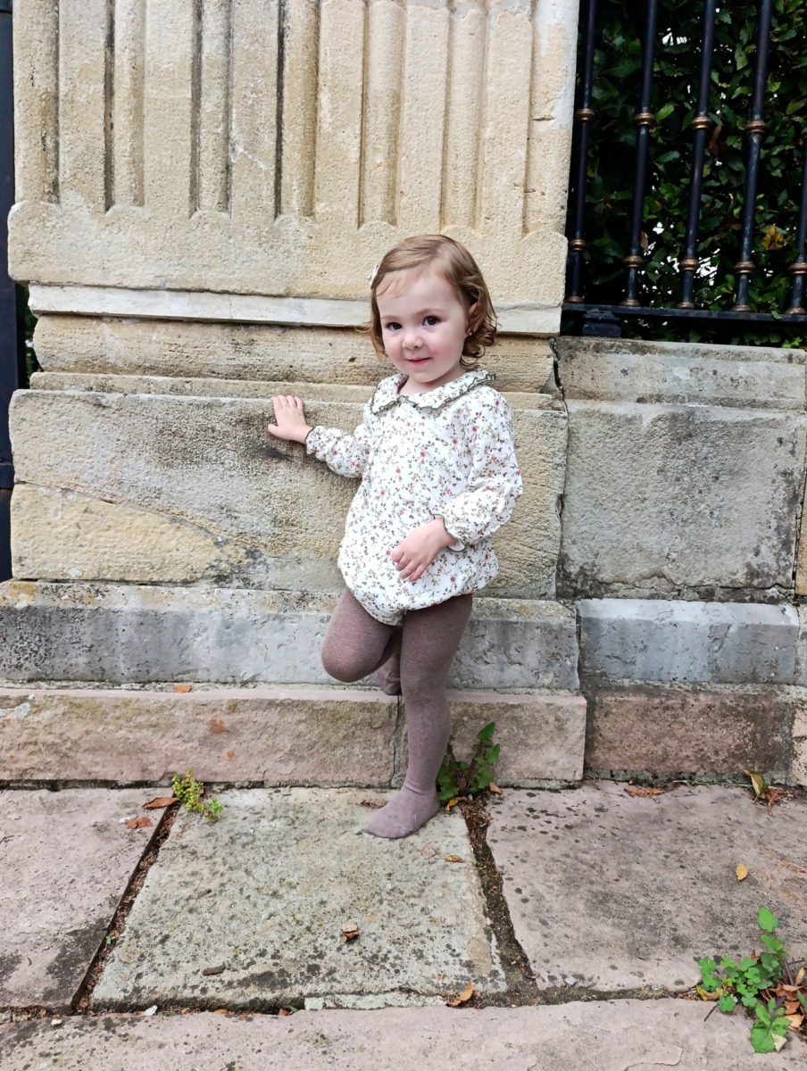 Niña pequeña con pelele de flores posando junto a una pared de piedra, moda infantil Sutier